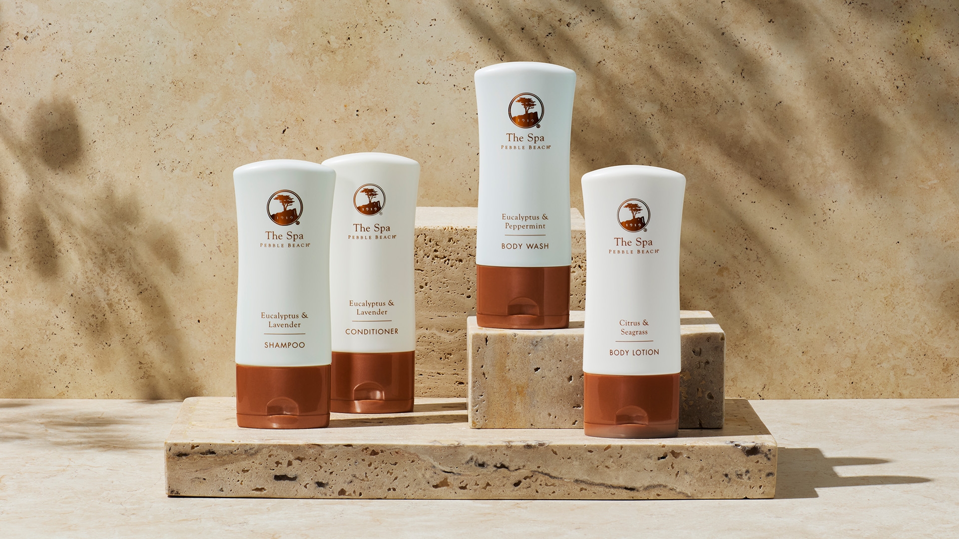 Four The Spa at Pebble Beach product bottles arranged on travertine blocks including shampoo, conditioner, body wash, and body lotion